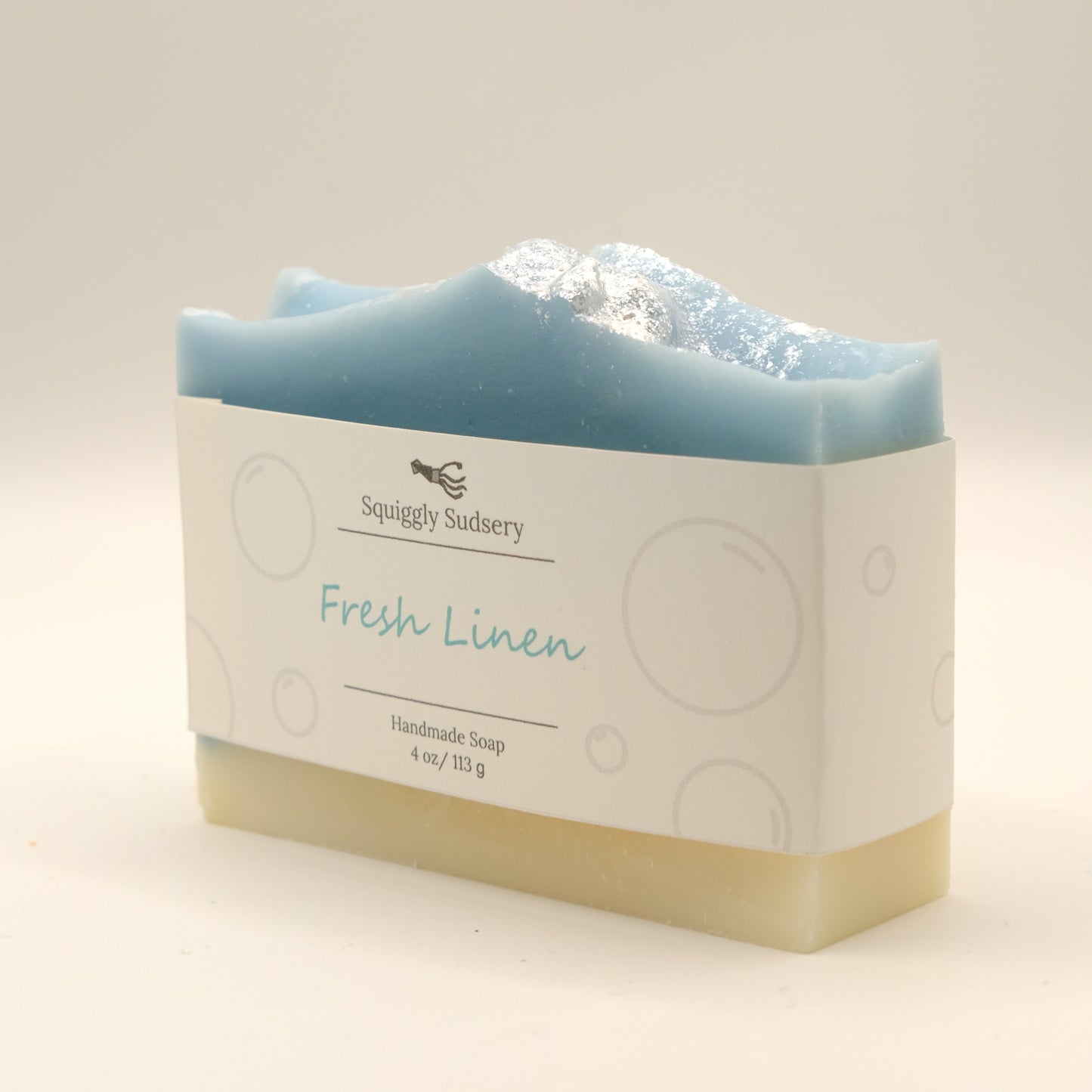 Blue and beige bar of soap with a white label 'Fresh Linen' by Squiggly Sudsery on a light beige background.