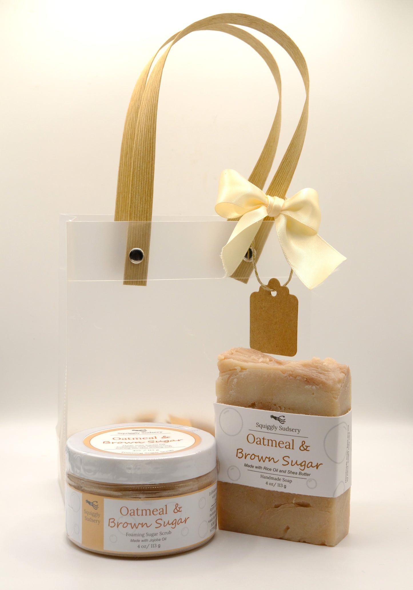 Jar of oatmeal and brown sugar foaming scrub and bar soap with a gift bag in the background on a white surface.