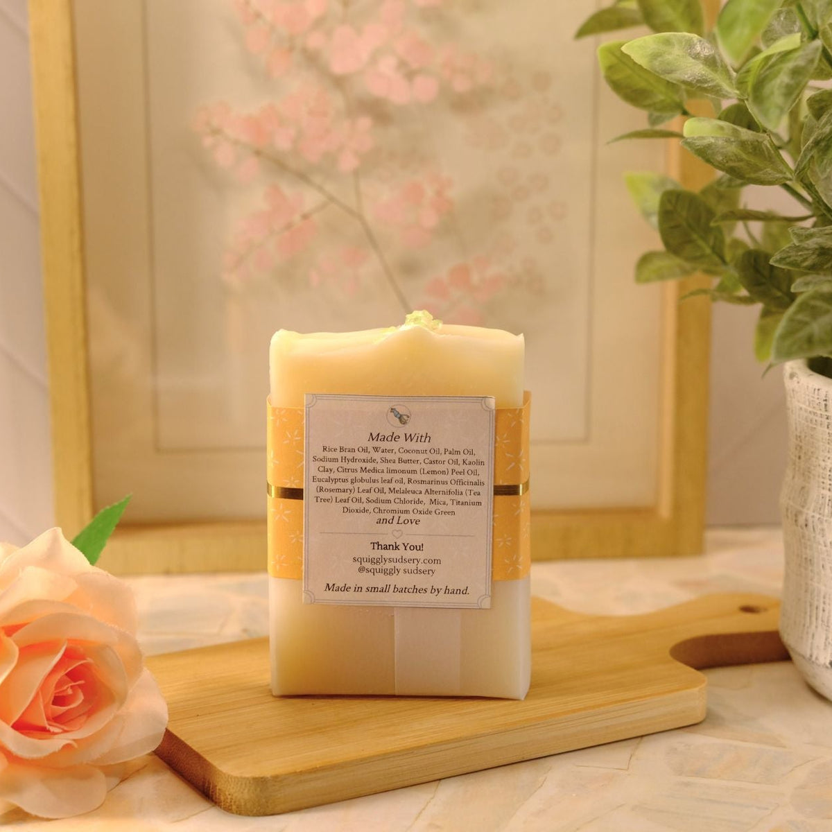 A handmade soap bar wrapped in a kraft paper band and a white paper label, displayed on a wooden cutting board with a peach rose and a potted plant in a styled setting.
