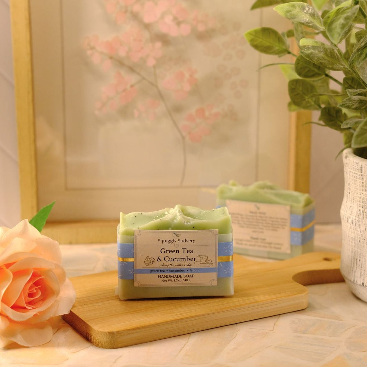 A handmade soap bar labeled 'Green Tea & Cucumber' sits on a wooden cutting board on a light patterned tablecloth, styled with a peach rose, a potted green plant, and a framed print in the background.