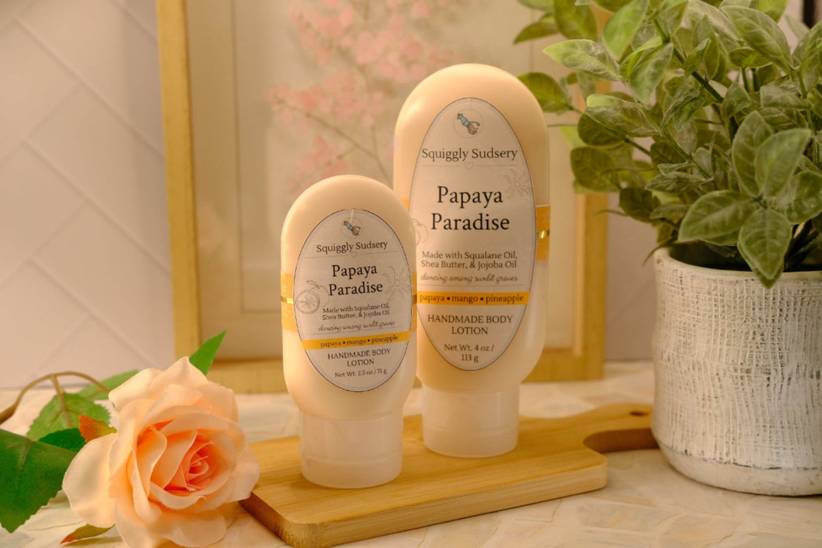 Two Squiggly Sudsery 'Papaya Paradise' lotions, displayed on a wooden block with a peach rose and a potted plant in the background.