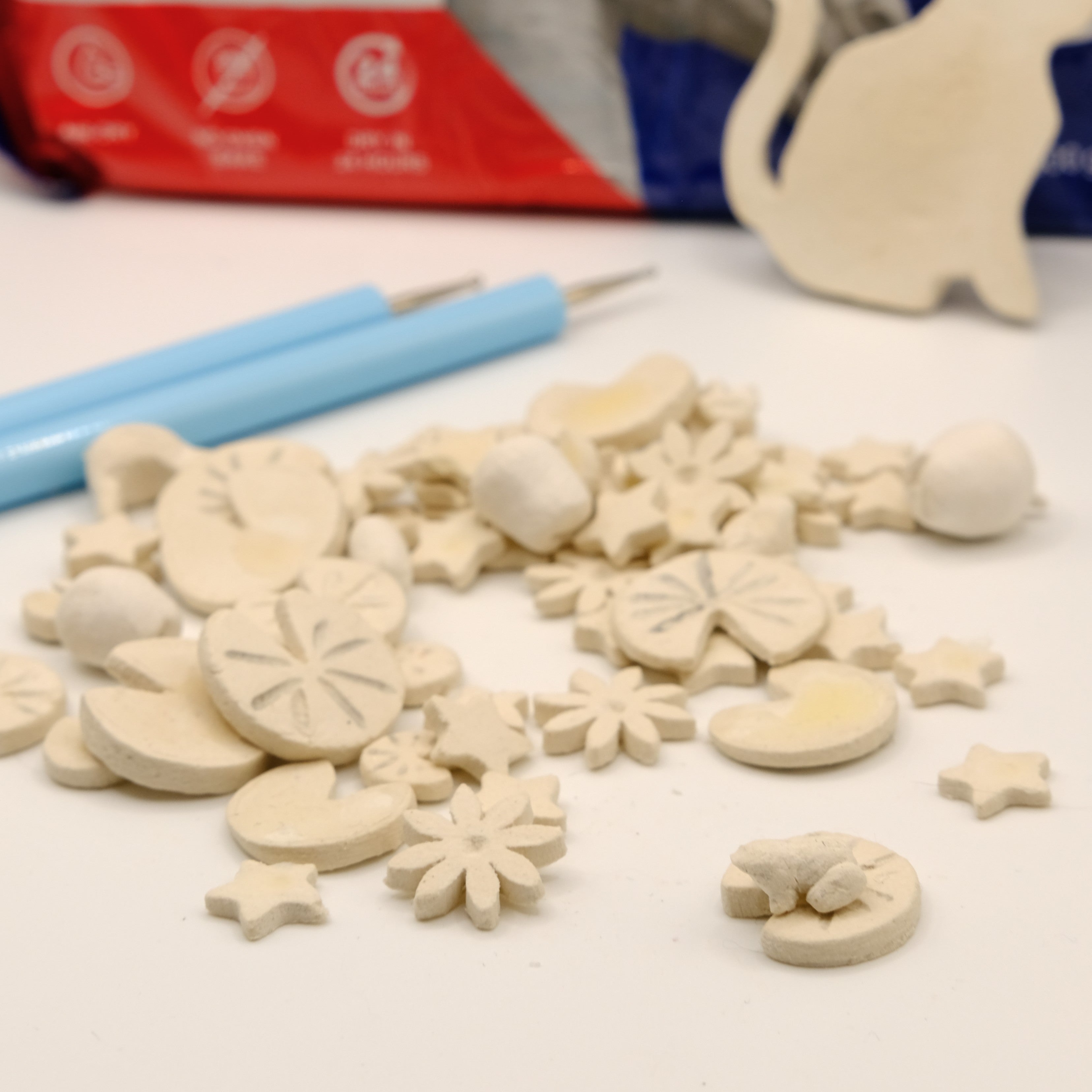 Small clay figures including a frog on a lily pad and small stars on a white surface with tools on the side and a red box in the background. 