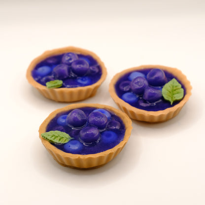 Three small tart-shaped soaps with blue fruit filling and mint leaves on a white background