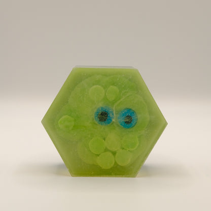 Hexagonal green soap bar embedded with green spheres and eye-shaped designs on a light gray background