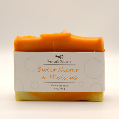 Orange and yellow bar of soap with 'Squiggly Sudsery' branding and 'Sweet Nectar & Hibiscus' description on a light background