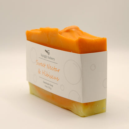 Three-quarter view of an orange and yellow bar of soap with 'Squiggly Sudsery' branding and 'Sweet Nectar & Hibiscus' description on a light background