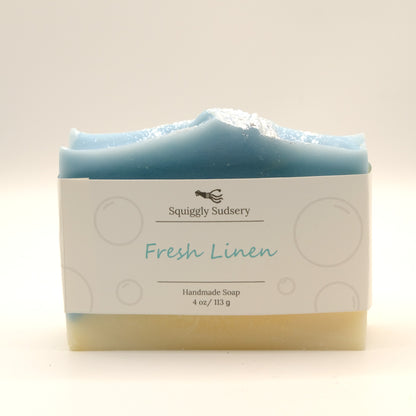 Blue and beige bar of soap with packaging labeled 'Squiggly Sudsery Fresh Linen' on a light background