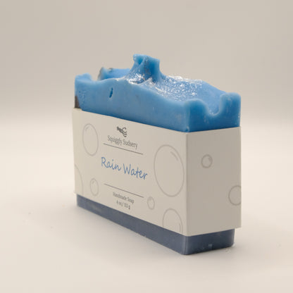 Blue soap bar with a white label 'Rain Water' with a textured top on a light gray background