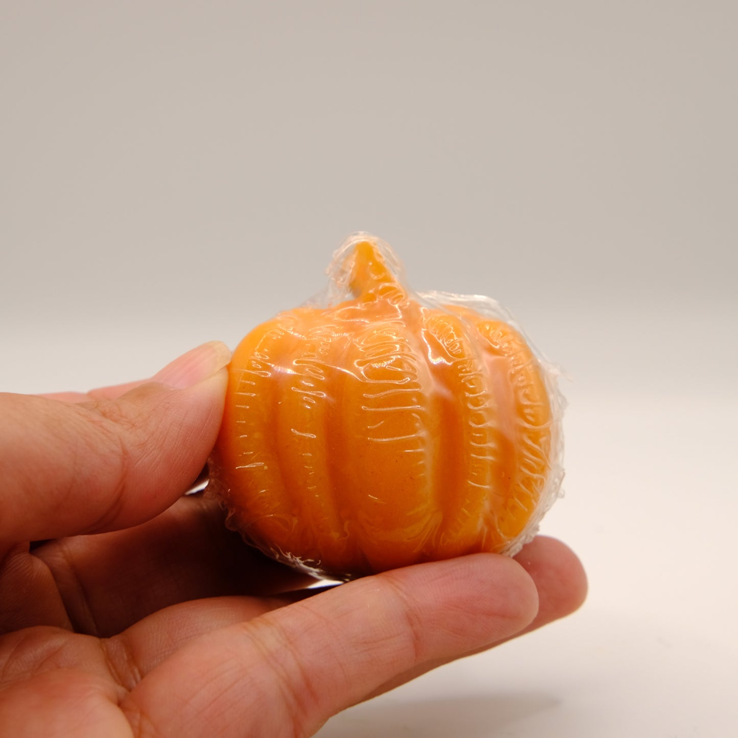Small orange pumpkin-shaped soap wrapped in plastic held by a hand against a plain background