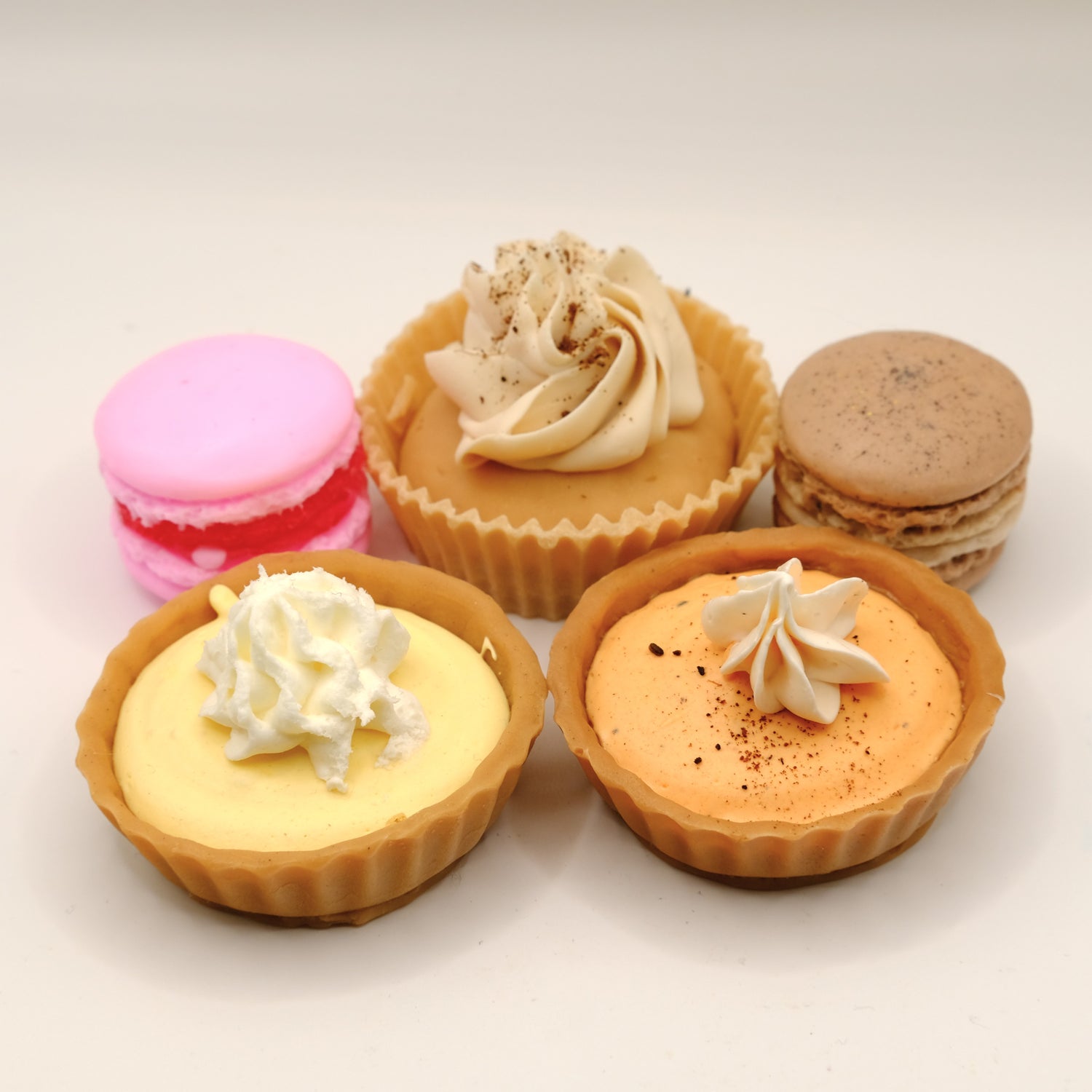 Three small tarts with whipped cream on top, surrounded by colorful macarons on a light background. All made with soap.