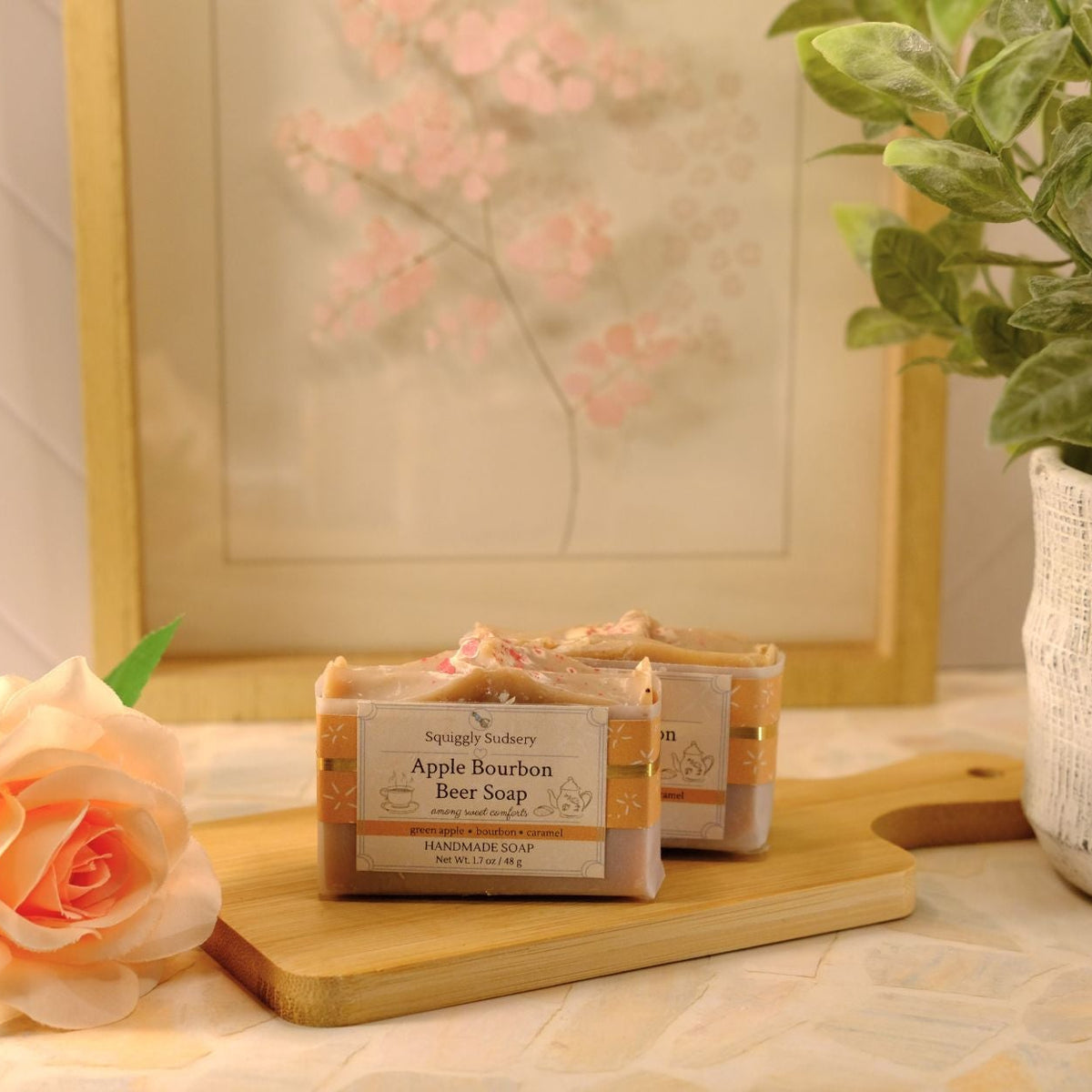 Two bars of soap labeled 'Apple Bourbon' on a wooden cutting board, styled with a peach rose and a potted plant against a warm, softly lit background.