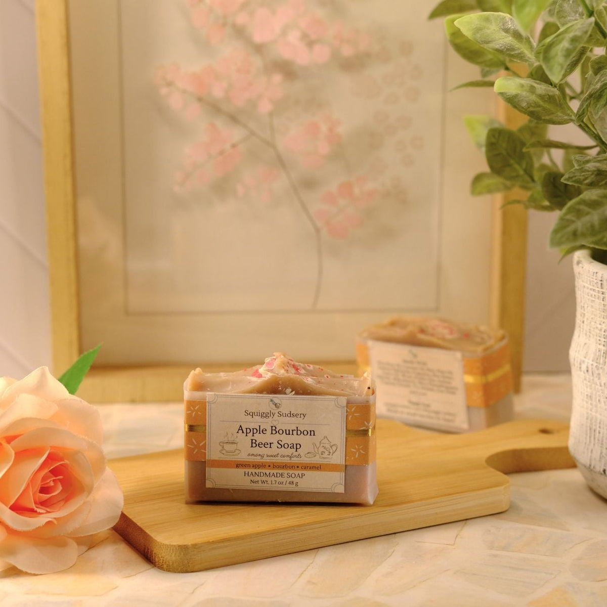 A bar of soap labeled 'Apple Bourbon Beer Soap' sits on a wooden cutting board on a table, styled with a peach rose, a potted green plant, and a framed print in the background.