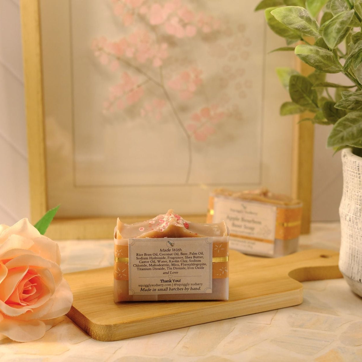 A handmade soap bar with a cream and gold label sits on a wooden cutting board, styled with a peach rose, a potted green plant, and a framed floral print in the background.