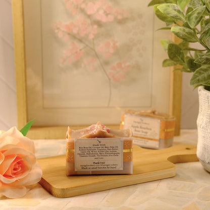 A handmade soap bar with a cream and gold label sits on a wooden cutting board, styled with a peach rose, a potted green plant, and a framed floral print in the background.