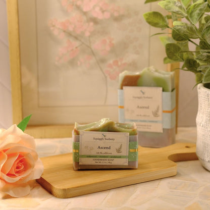 A bar of soap labeled 'Ascend' by Squiggly Sudsery on a wooden cutting board on a tiled countertop, styled with a peach rose, a potted plant, and a framed print in the background.