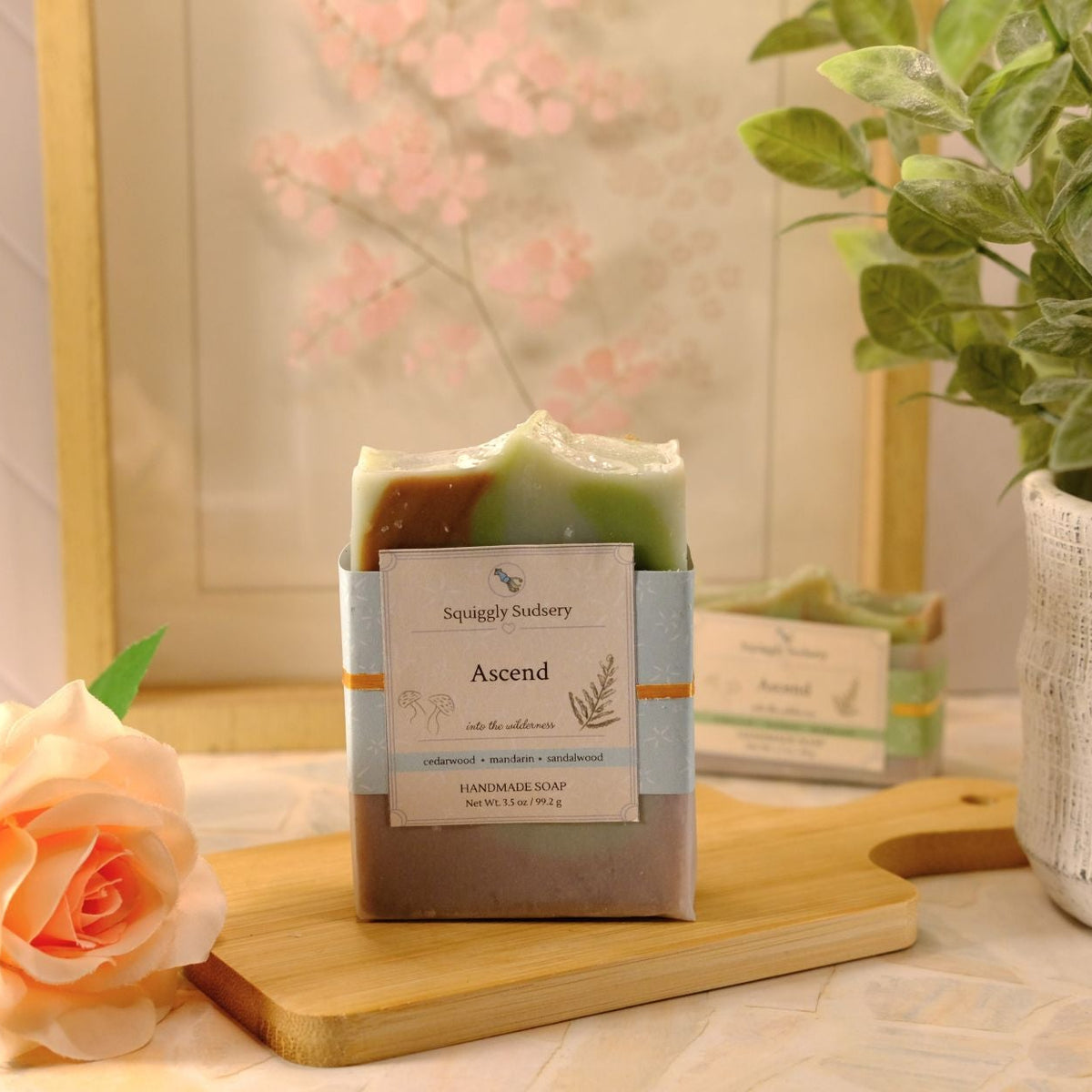 A packaged handmade soap bar labeled 'Ascend' by Squiggly Sudsery, displayed on a wooden board with a peach rose and a potted plant in a softly styled indoor setting.
