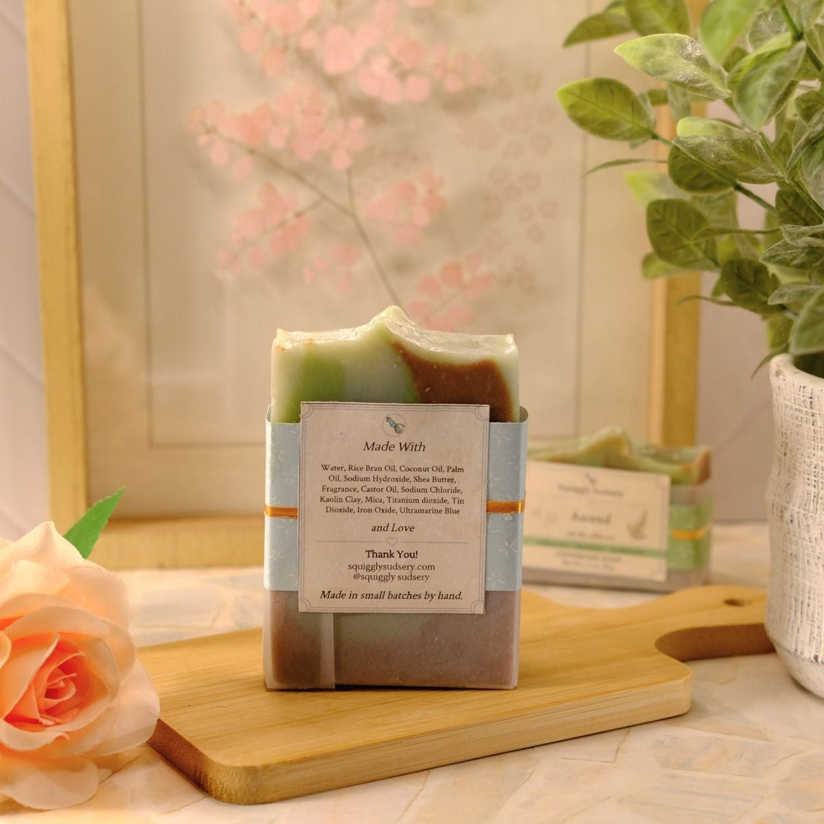 A handmade soap bar wrapped in kraft paper and a paper label, displayed on a wooden cutting board with a peach rose and a potted plant in the background.