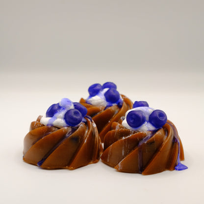 Three bundt cake-shaped soaps with blueberries, blue drizzle and white filling on a light gray background