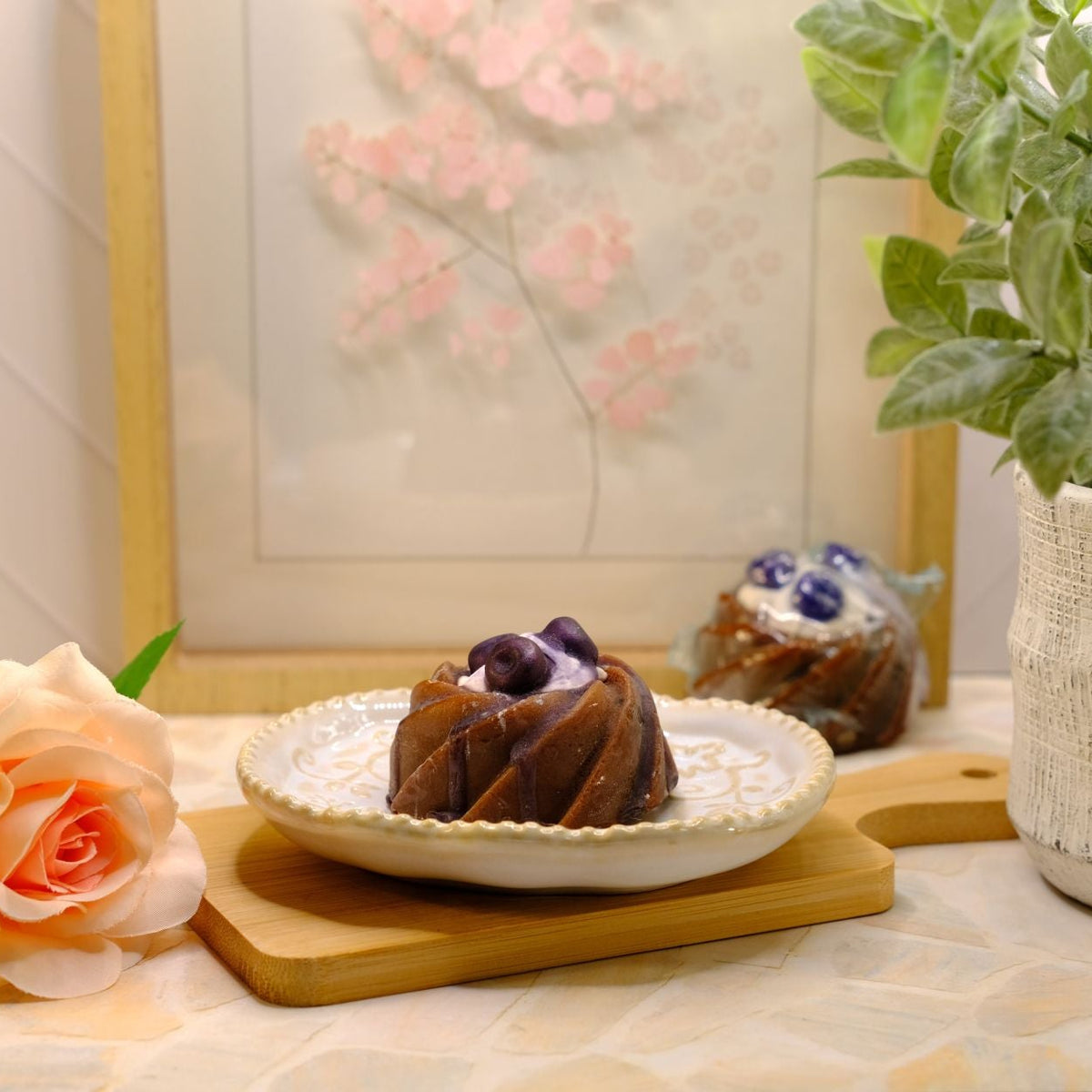 A small blueberry cake soap on a white plate with purple berries, styled on a wooden board beside a peach rose, a potted green plant, and a framed floral print in a warm indoor setting.