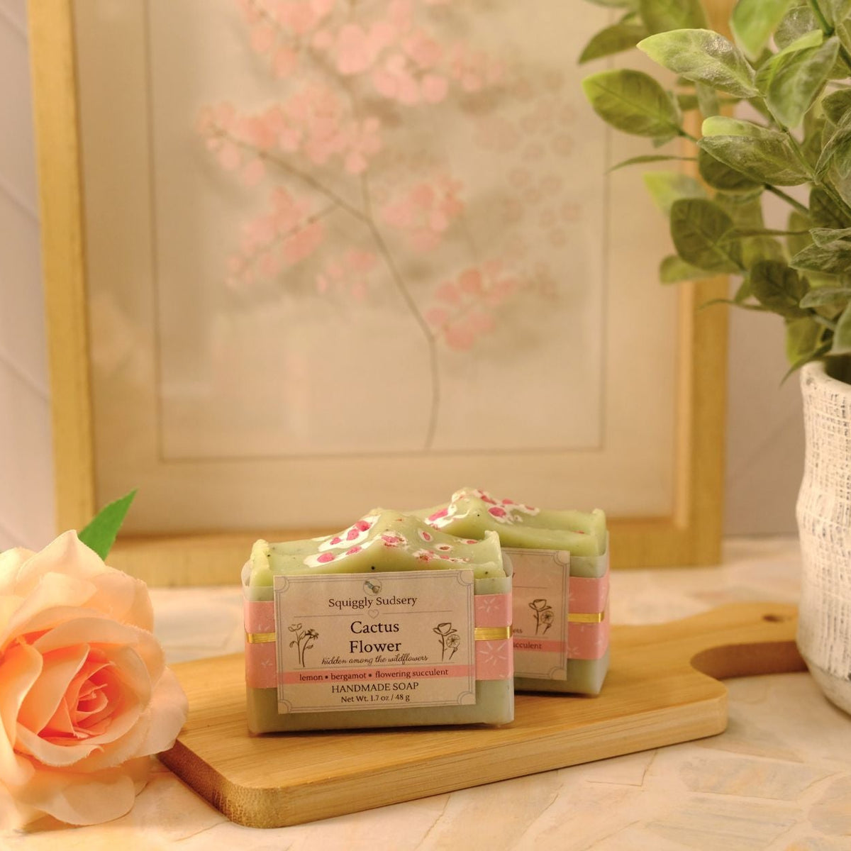 Two wrapped bars of soap labeled 'Cactus Flower' by Squiggly Sudsery, displayed on a wooden board on a tiled countertop, with a peach rose, a potted green plant, and a framed print in the background.