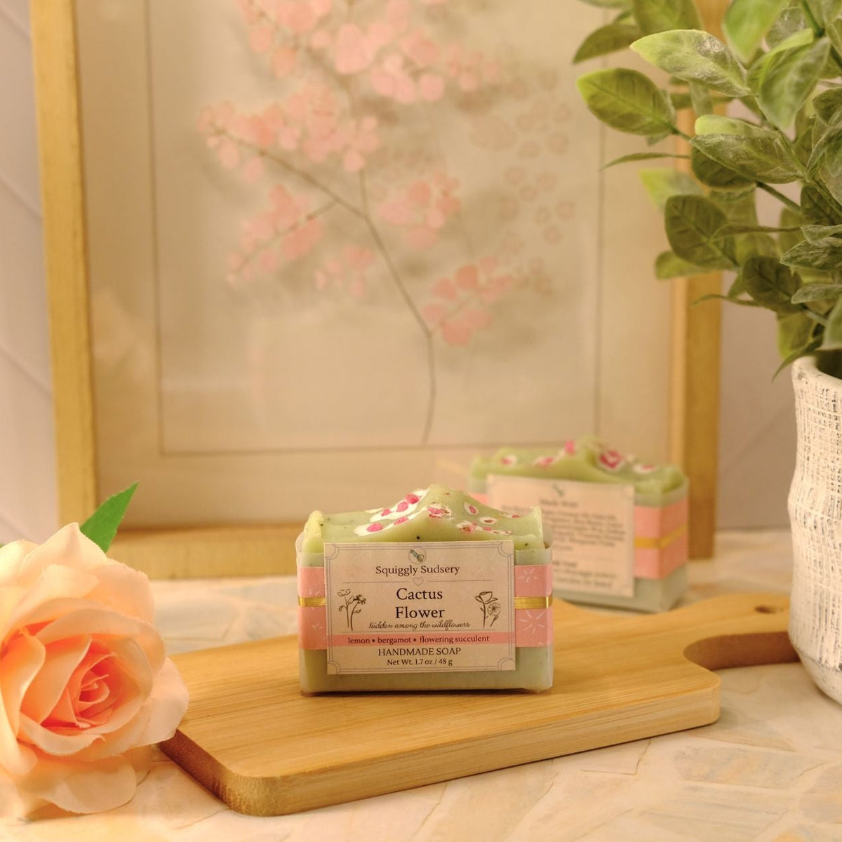A bar of soap labeled 'Cactus Flower' by Squiggly Sudsery, sits on a wooden cutting board on a patterned tablecloth, styled with a peach rose, a potted plant, and a framed print in the background.