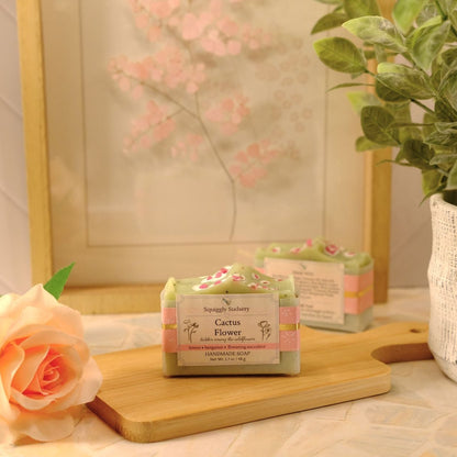 A bar of soap labeled 'Cactus Flower' by Squiggly Sudsery, sits on a wooden cutting board on a patterned tablecloth, styled with a peach rose, a potted plant, and a framed print in the background.