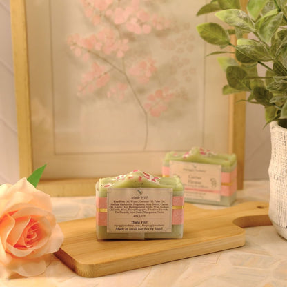 A small rectangular bar of soap with a pastel green and pink wrapper sits on a wooden cutting board on a tabletop, styled with a peach rose, a potted green plant, and a framed floral print in the background.