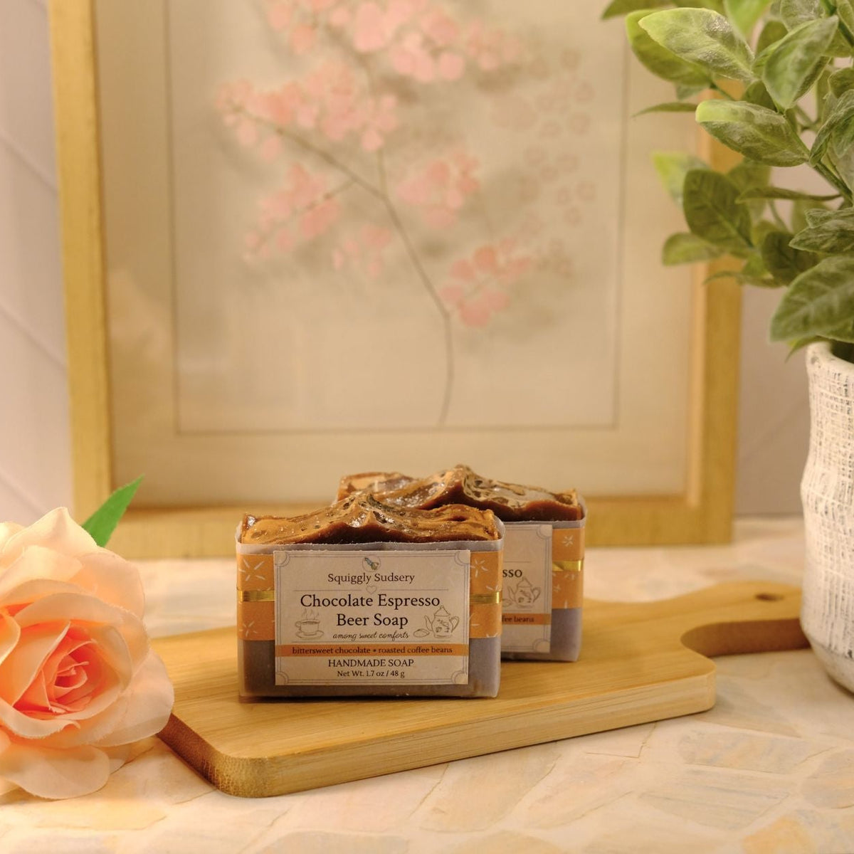 A bar of soap labeled 'Chocolate Espresso Beer Soap' sits on a wooden cutting board on a patterned tablecloth, styled with a peach rose, a potted green plant, and a framed picture in the background.
