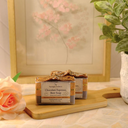 A bar of soap labeled 'Chocolate Espresso Beer Soap' sits on a wooden cutting board on a patterned tablecloth, styled with a peach rose, a potted green plant, and a framed picture in the background.