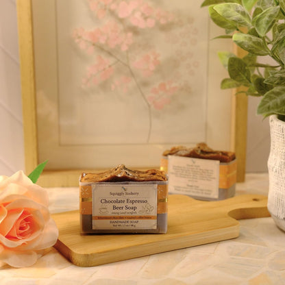 Two artisanal bars of soap labeled 'Chocolate Espresso Beer Soap' displayed on a wooden cutting board, styled with a peach rose, a potted green plant, and a framed print in a warm, soft-focus setting.