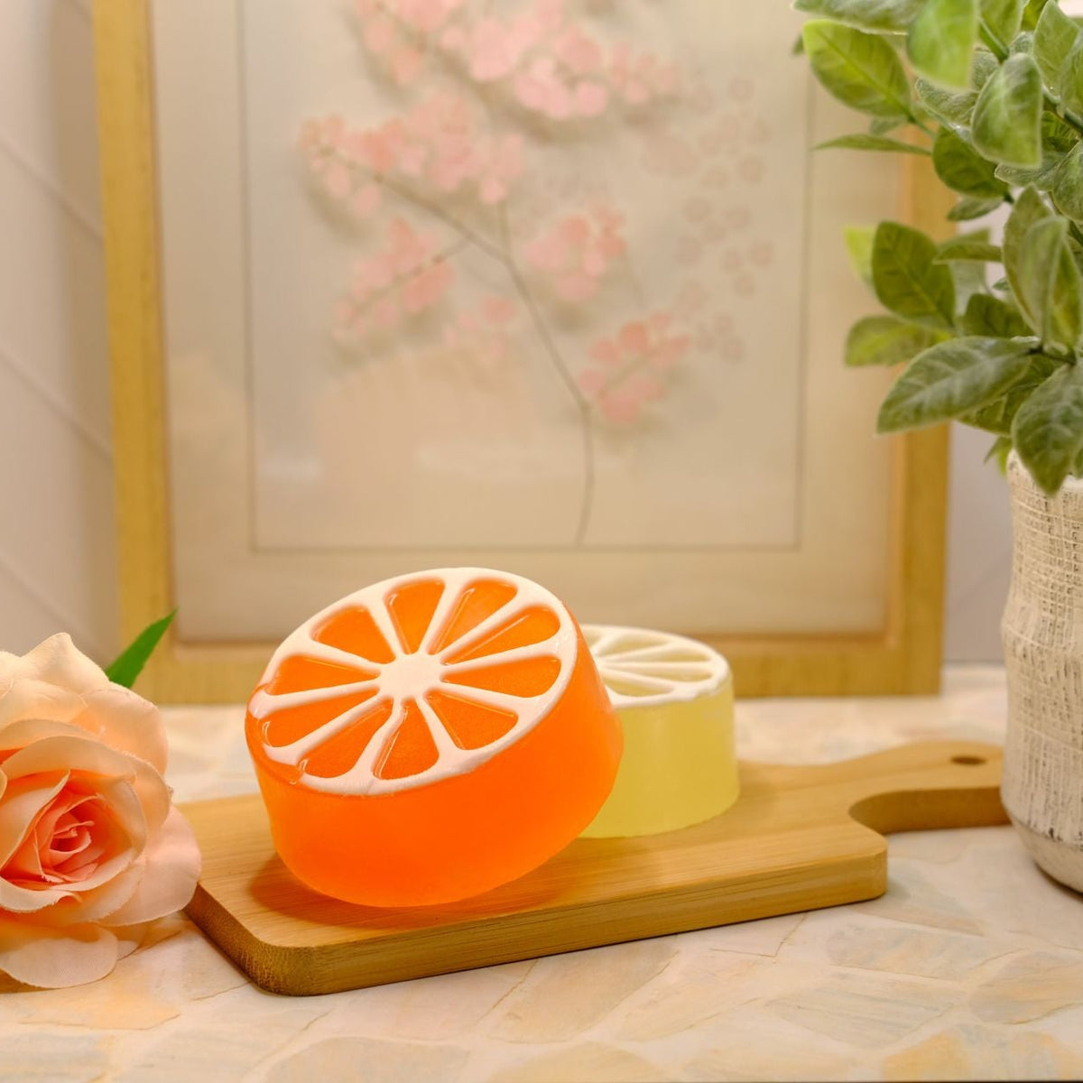 An orange and yellow citrus slice-shaped soap set on a wooden cutting board, styled with a peach rose and a potted plant in a warm indoor setting.