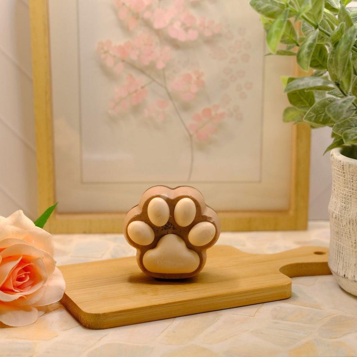A small, brown paw-print shaped soap sits on a wooden cutting board, styled with a peach rose, a potted green plant, and a framed print in the background.