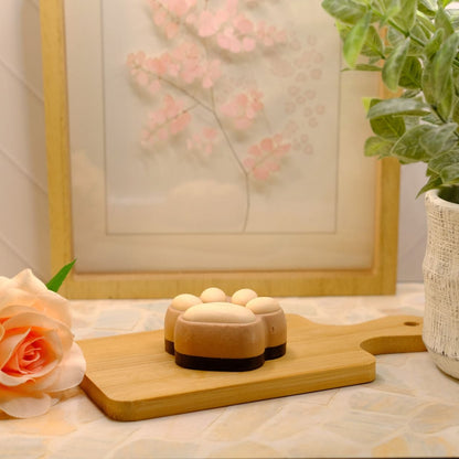 A brown paw-shaped object with small round beige bumps on top, displayed on a wooden cutting board in a styled tabletop setting with a peach rose, framed floral artwork, and a potted green plant.