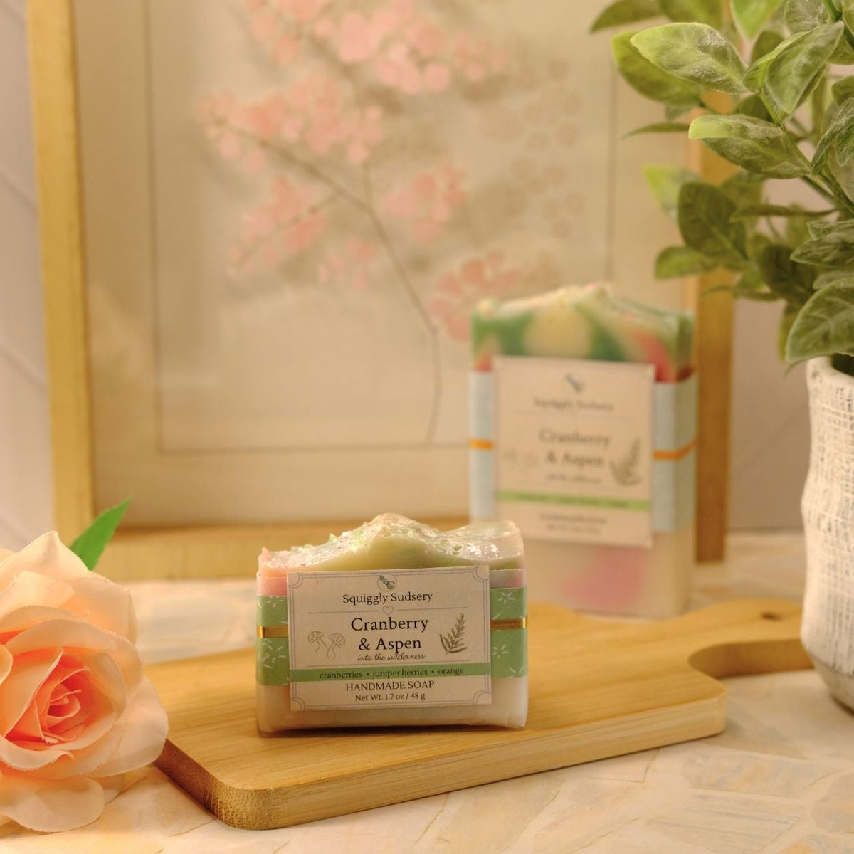 A bar of soap labeled Cranberry & Aspen by Squiggly Sudsery sits on a wooden cutting board, styled with a peach rose, a potted green plant, and a framed floral print in a warm, soft setting.