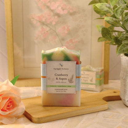 A handmade soap bar labeled 'Cranberry & Aspen' by Squiggly Sudsery, displayed on a wooden cutting board. The soap has a green, white, and pink marbled pattern.