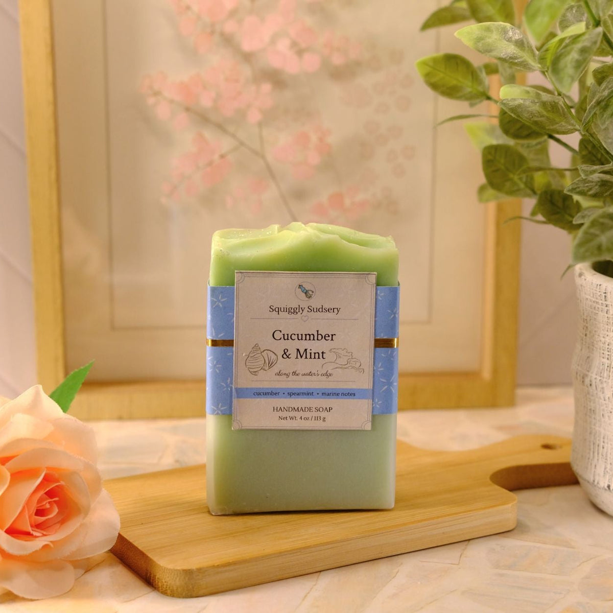 A handmade soap bar labeled 'Cucumber & Mint' by Squiggly Sudsery, standing on a wooden cutting board. The scene is styled with a peach rose, a potted green plant, and a framed picture in the background.