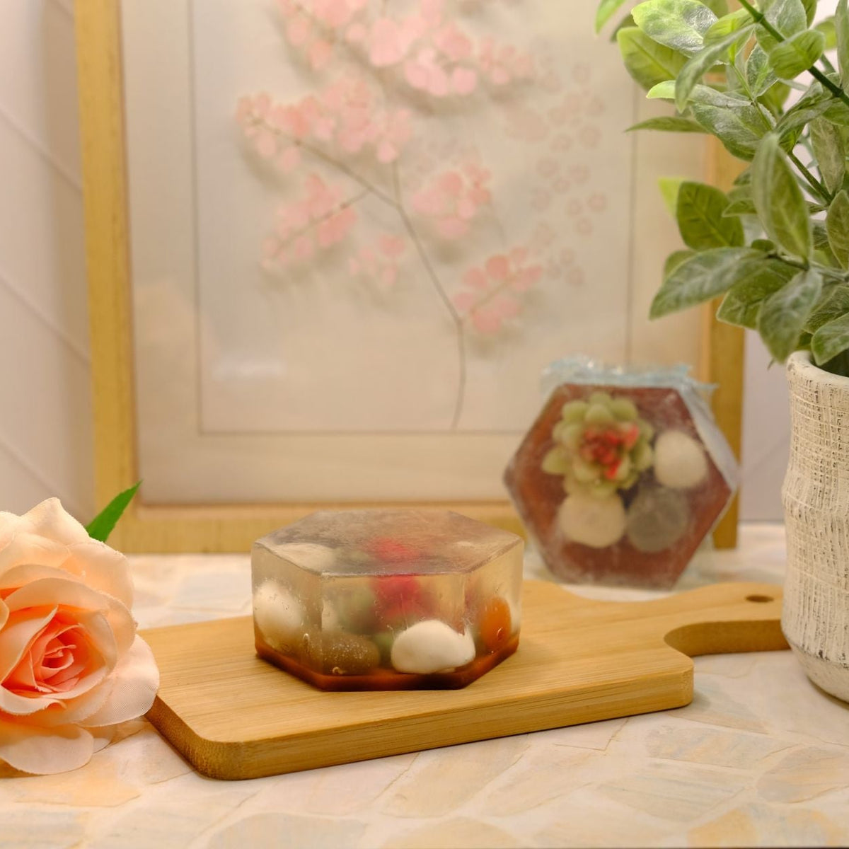 A small hexagonal soap containing colorful decorative stones and a tiny succulent, displayed on a wooden cutting board next to a peach rose and a potted green plant.