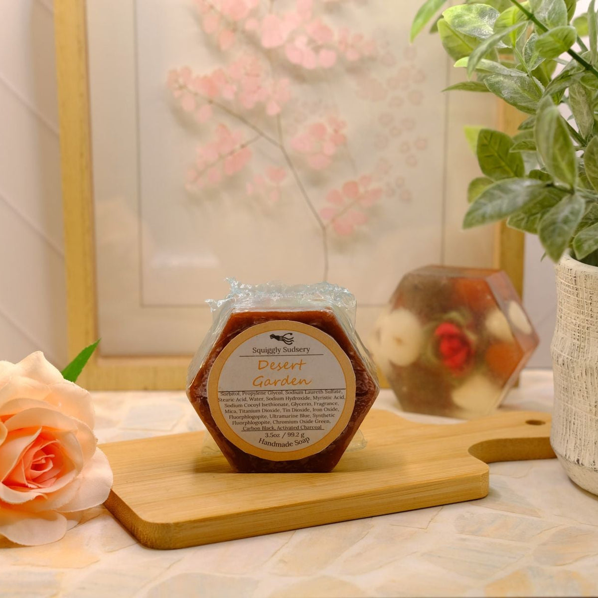 A hexagonal bar of Desert Garden displayed on a wooden cutting board, styled with a peach rose, a potted green plant, and a framed floral print in the background.