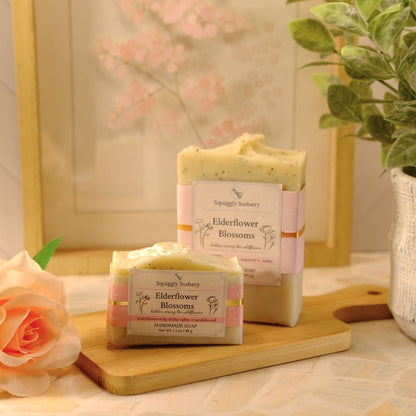Two handmade soap bars labeled Elderflower Blossom by Squiggly Sudsery, styled on a wooden cutting board with a peach rose and a potted plant in a warm, soft-focus setting.
