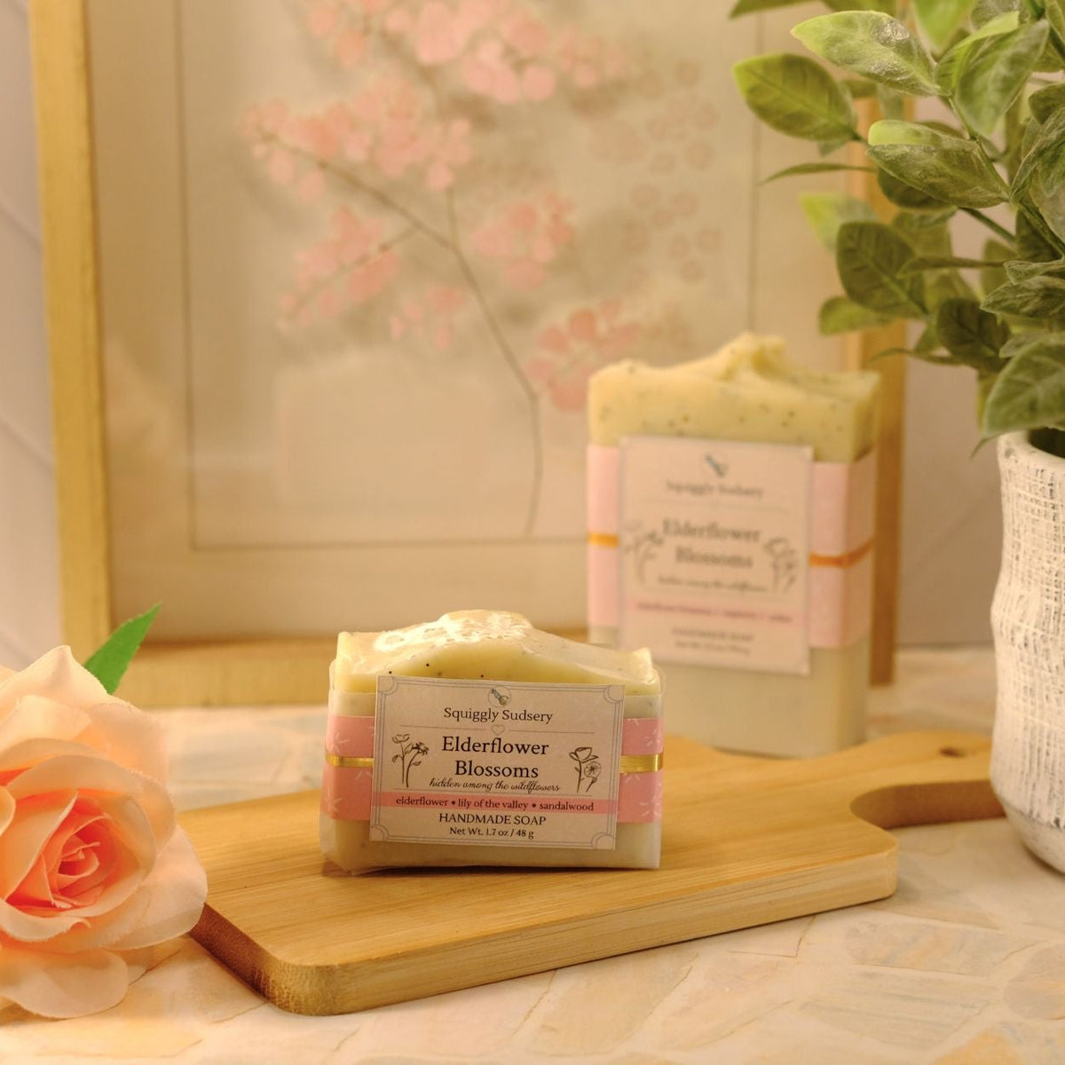 Two handmade soap bars from the Squiggly Sudsery styled on a wooden cutting board with a peach rose and a potted plant in a warm, soft-focus setting.