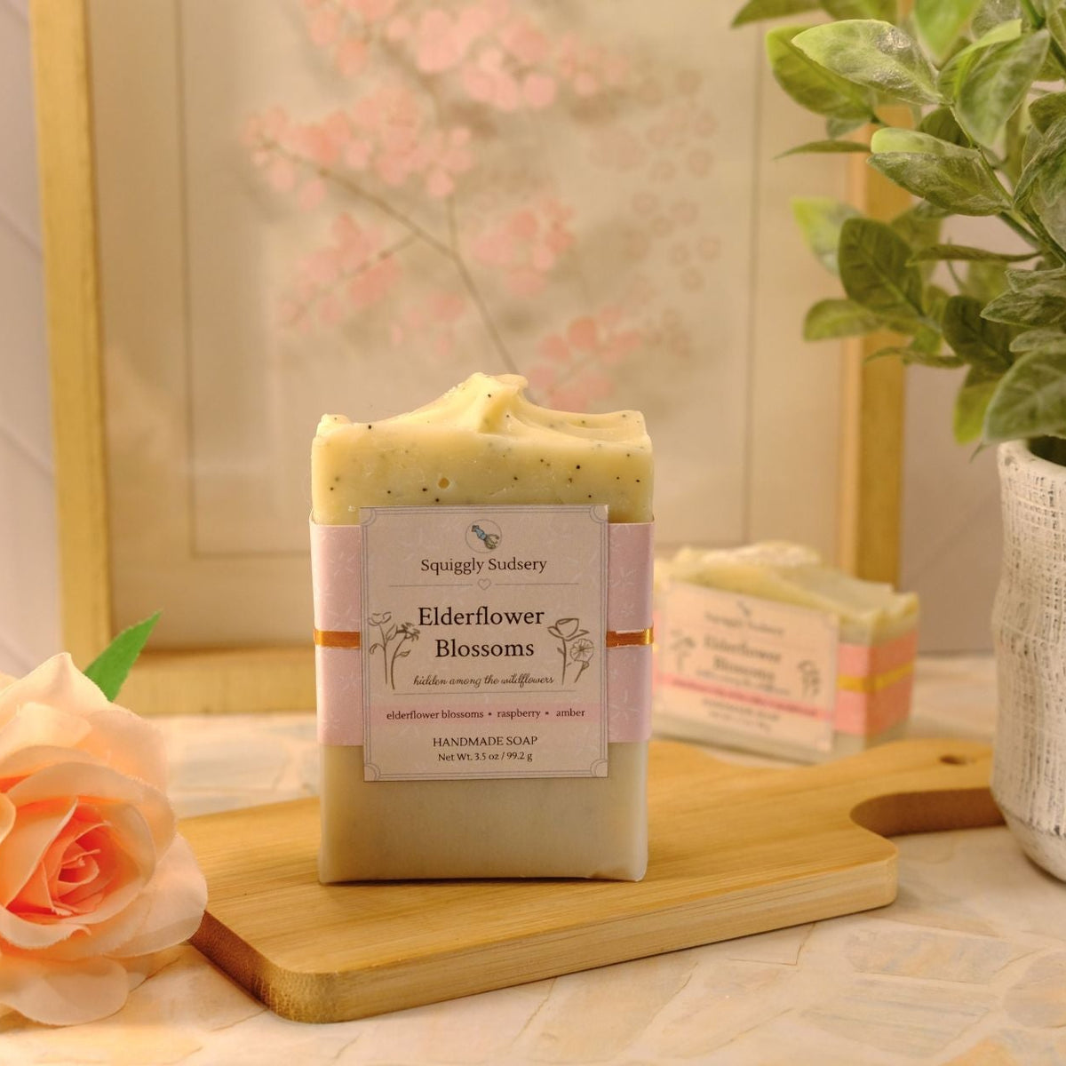 A handmade rectangular bar of soap labeled 'Elderflower Blossoms' by Squiggly Sudsery, displayed on a wooden cutting board with a peach rose and a potted plant in a styled setting.
