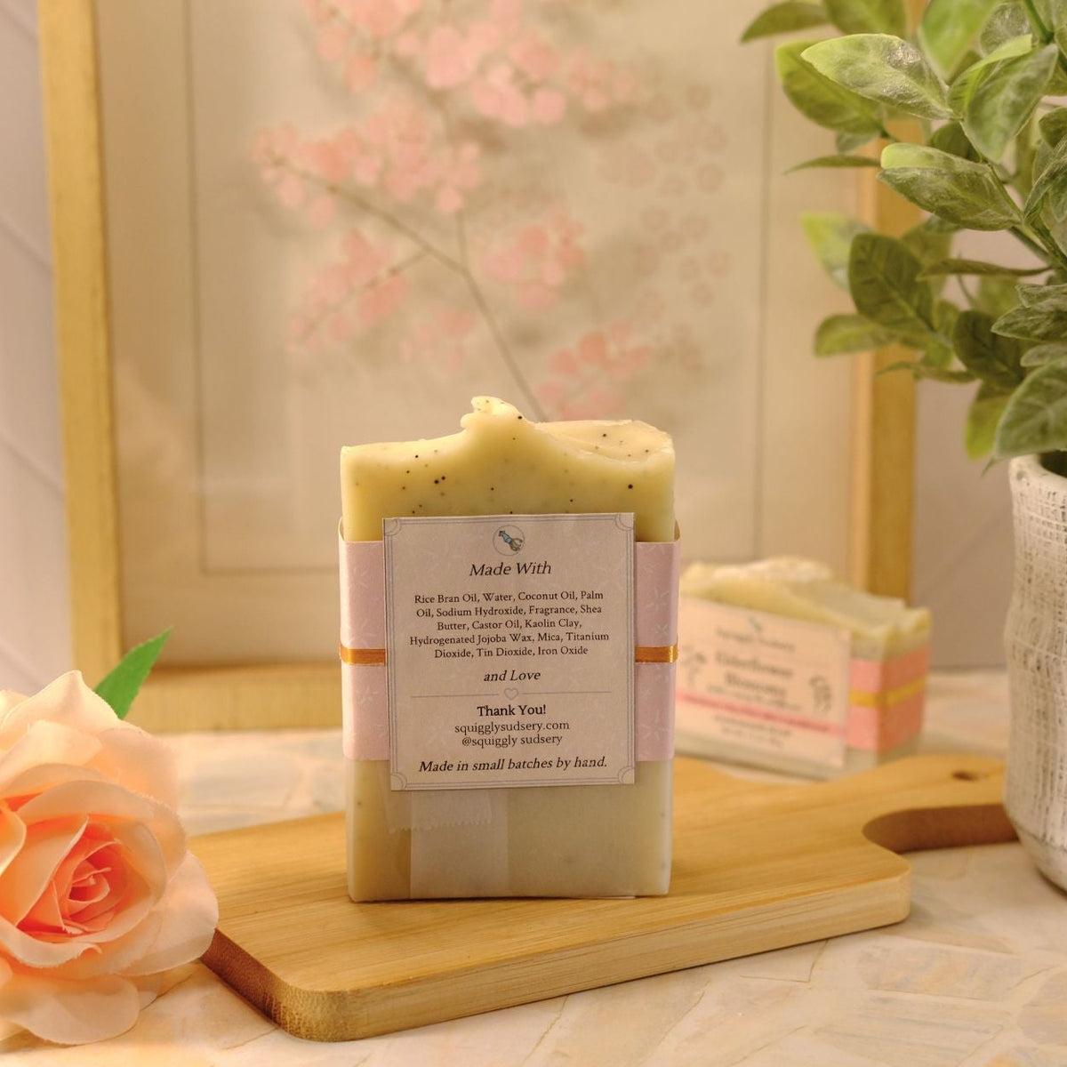 A handmade soap bar with a white paper label sits on a wooden cutting board, styled with a peach rose, a potted green plant, and a blurred soap bar in the background.