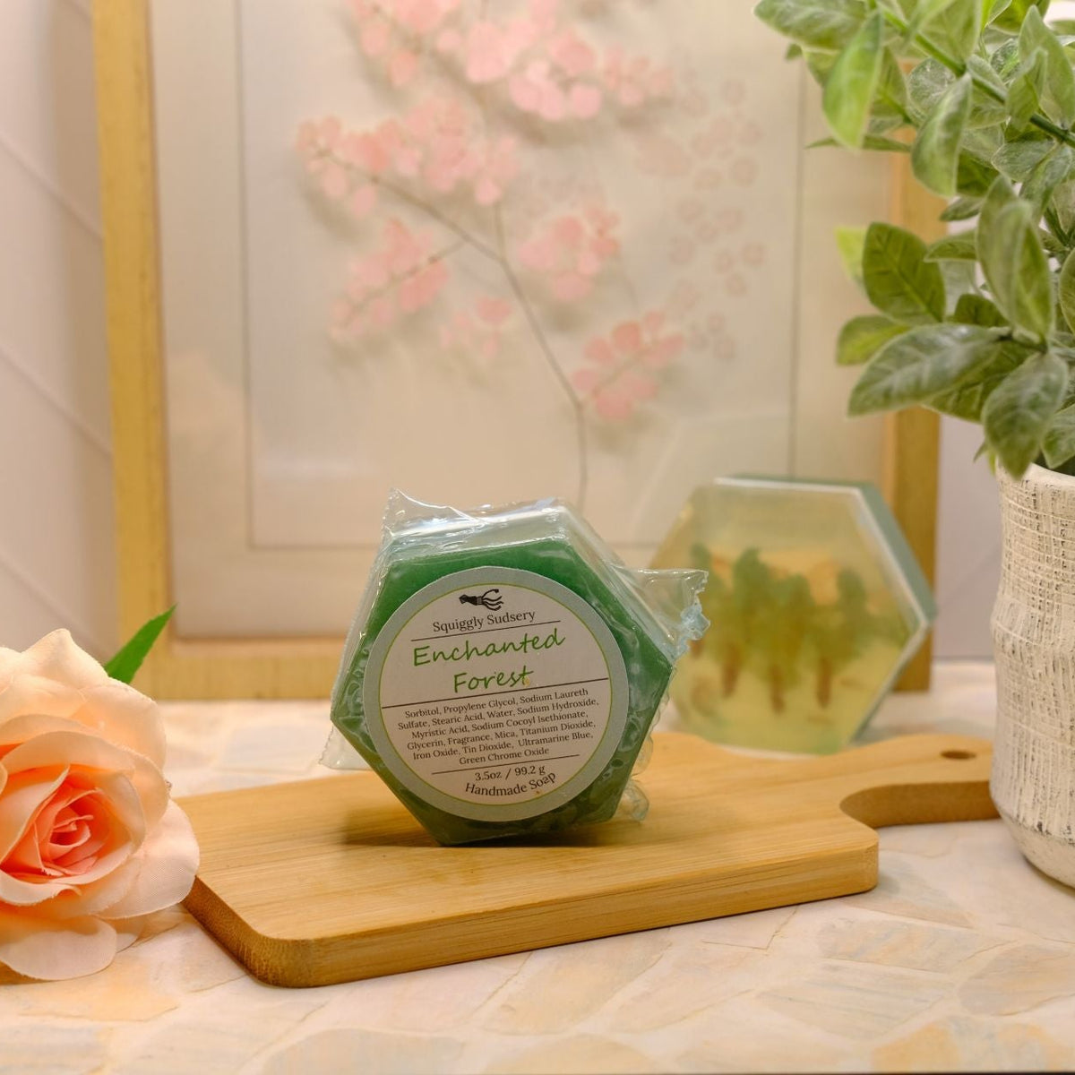 A hexagonal green soap bar labeled 'Enchanted Forest' sits on a wooden cutting board on a patterned tablecloth, styled with a peach rose, a potted green plant, and a framed print in the background.
