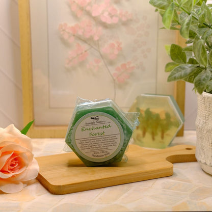 A hexagonal green soap bar labeled 'Enchanted Forest' sits on a wooden cutting board on a patterned tablecloth, styled with a peach rose, a potted green plant, and a framed print in the background.