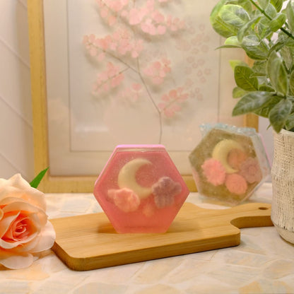 A pink hexagonal handmade soap with a crescent moon and flower design sits on a wooden cutting board on a table, styled with a peach rose, a potted green plant, and a framed floral print in the background.
