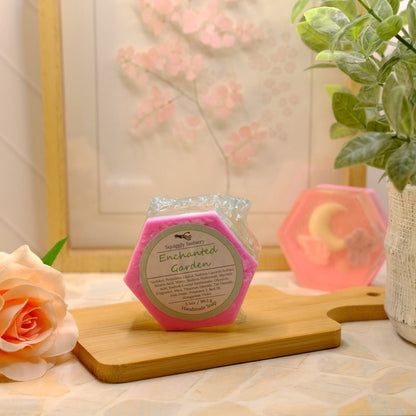 A hexagonal pink soap labeled 'Enchanted Garden' sits on a wooden cutting board on a tabletop, styled with a peach rose, a potted green plant, and another pink hexagonal candle in the background.