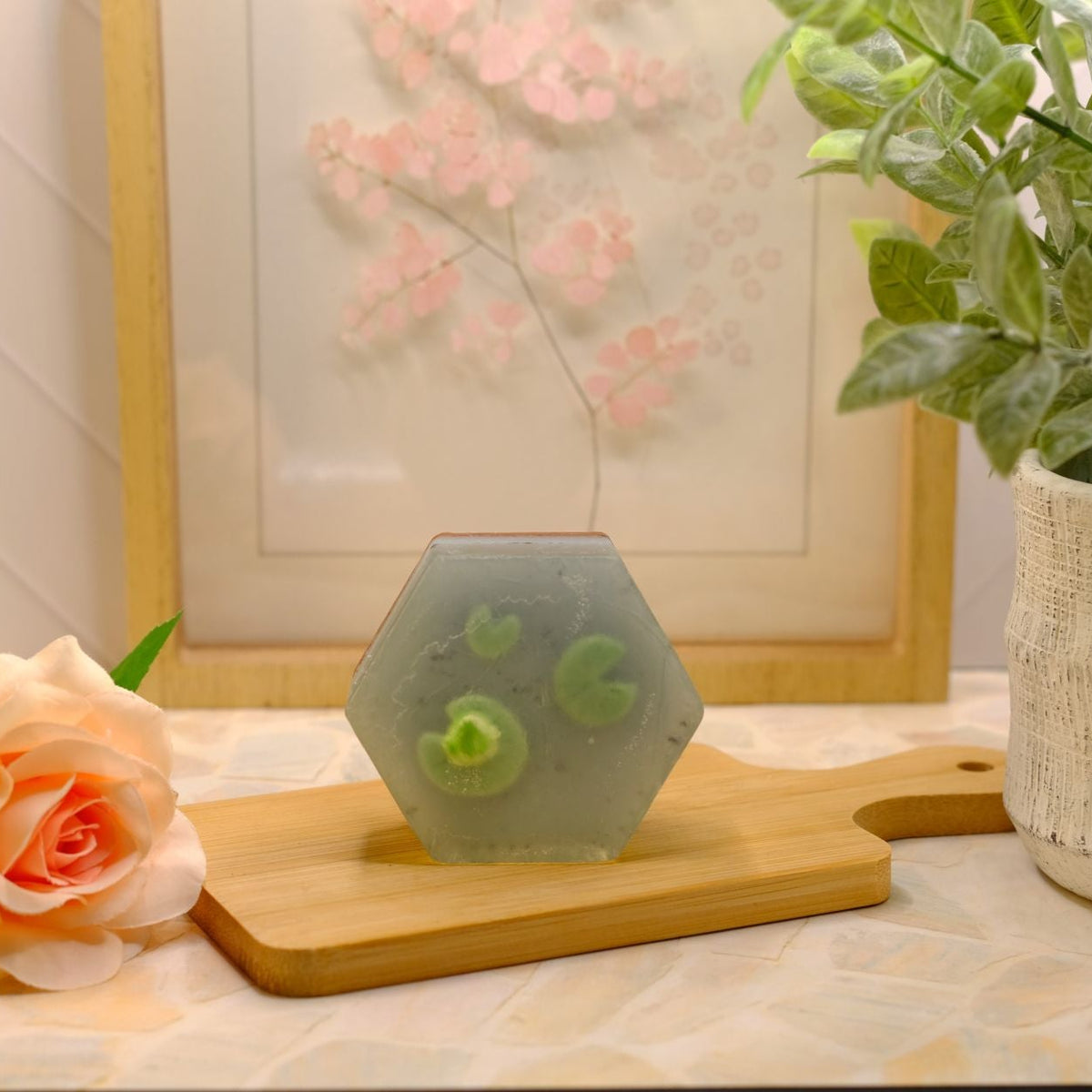 Two hexagonal translucent blue soap bars with three green lily pads and a miniature green soap frog, displayed on a wooden cutting board on a tiled countertop, styled with a peach rose and a potted plant.
