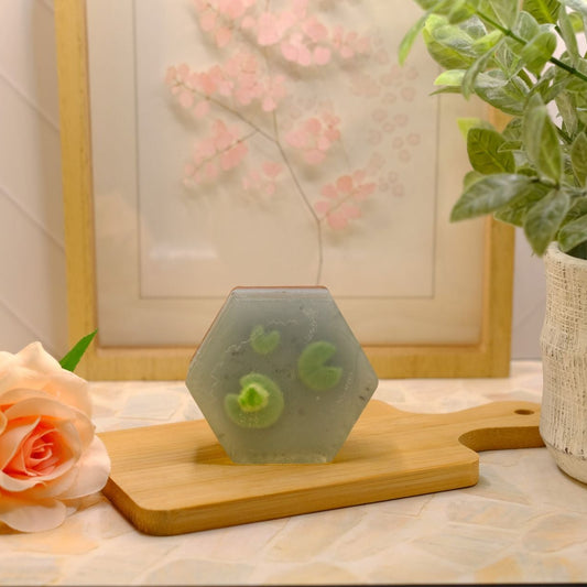 Two hexagonal translucent blue soap bars with three green lily pads and a miniature green soap frog, displayed on a wooden cutting board on a tiled countertop, styled with a peach rose and a potted plant.