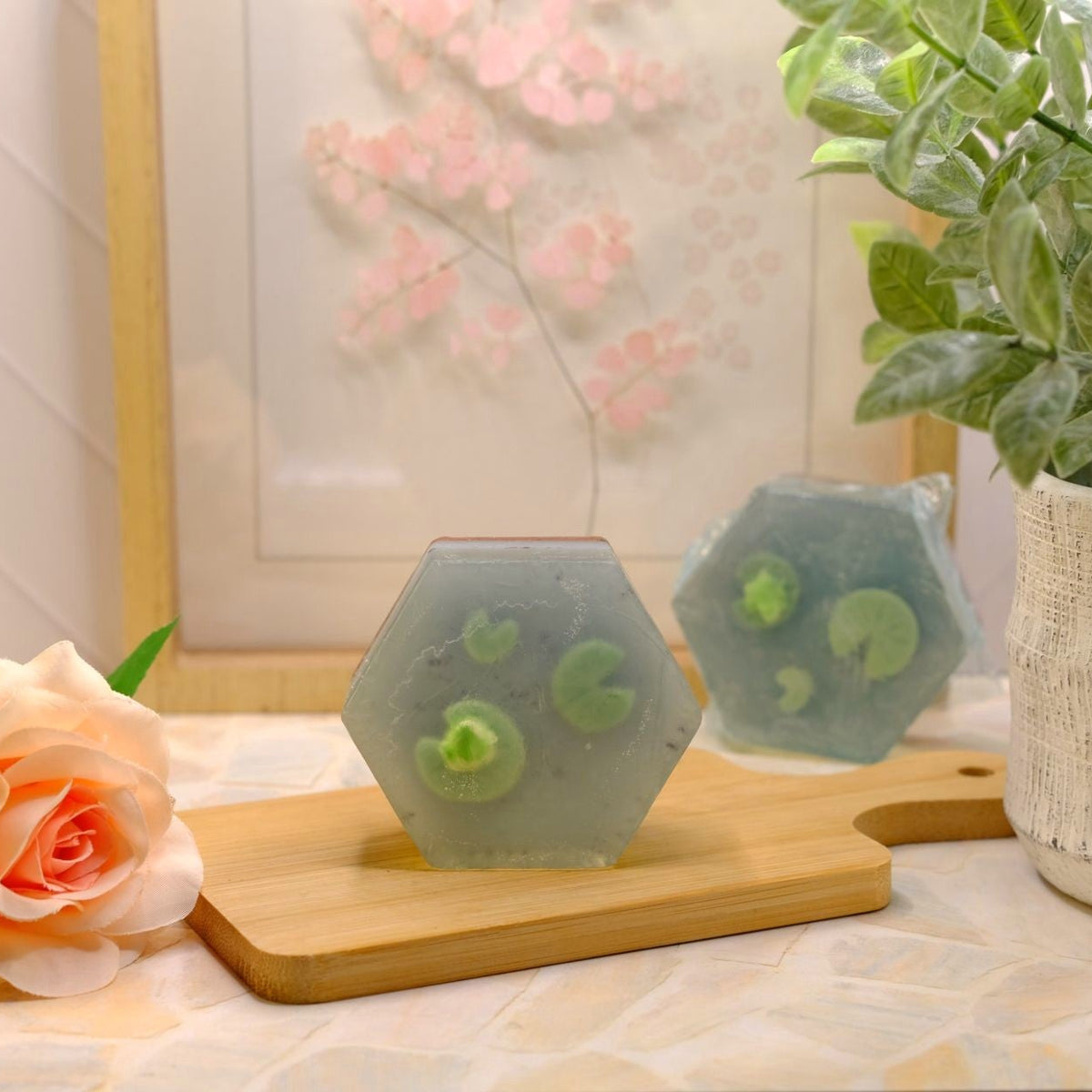 Two hexagonal translucent green soap bars with circular inclusions, displayed on a wooden cutting board on a tiled countertop, styled with a peach rose and a potted plant.