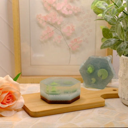 A hexagonal handmade soap bar with green botanical inclusions sits on a wooden cutting board, styled with a peach rose, a textured planter, and a framed floral print in the background.
